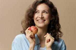 heiter lockig süß Dame im Jeans beiläufig Hemd mit Apfel in der Nähe von Wange sieht aus beiseite posieren isoliert auf Beige Pastell- Hintergrund. gesund Lebensmittel. natürlich umweltfreundlich Produkte Konzept. Kopieren Raum foto