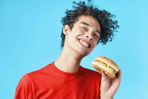 Kerl mit lockig Haar im rot T-Shirt Hamburger schnell Essen Blau Hintergrund foto