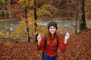 froh Frau im ein Hut Sweatshirt mit ein Rucksack auf ihr zurück Gesten mit ihr Hände im ein Park im Natur foto