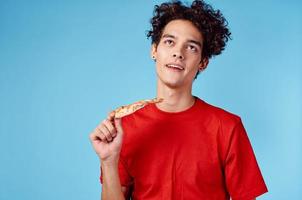 Mann halten ein Scheibe von Pizza im seine Hand und lockig Haar rot T-Shirt Blau Hintergrund foto