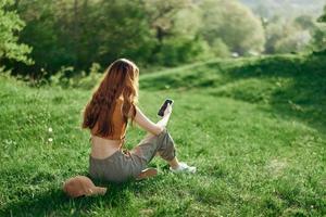 oben Aussicht von ein Frau im ein Orange oben und Grün Hose Sitzung auf das Sommer- Grün Gras mit ihr zurück zu das Kamera mit ihr Telefon, ein jung freiberuflich Studenten Konzept von Arbeit und Freizeit foto