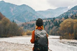 Frau steht auf das Fluss Bank und sieht aus beim das Berge im das Entfernung Landschaft Herbst Rucksack Tourismus Modell- foto