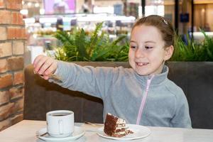 lächelnd wenig Mädchen im ein Cafe beim das Tabelle gießt Zucker in ein Tasse Nächster zu ein Teller mit Kuchen foto