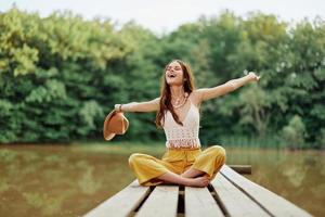 Hippie Öko-Aktivist Frau Reisender sitzt auf ein Brücke durch ein See mit ihr Waffen ausgestreckt mit ein Hut und lächelnd Mit freundlichen Grüßen foto