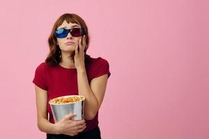 verwirrt konzentriert süß Rothaarige Dame im rot T-Shirt 3d Brille Essen Popcorn Uhr Film posieren isoliert auf Über Rosa Studio Hintergrund. Kopieren Raum Banner. Mode Kino Konzept. Unterhaltung Angebot foto