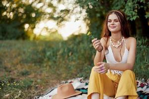 Mädchen gekleidet wie ein Hippie Öko entspannend im das Park, Sitzung auf ein Decke im das Sonnenuntergang, entspannt Lebensstil foto