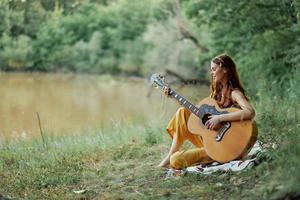 Hippie Frau spielen Gitarre lächelnd und Singen Lieder im Natur Sitzung auf ein Plaid durch das See im das Abend im das Strahlen von das Rahmen Sonne. ein Lebensstil im Harmonie mit das Körper und Natur foto