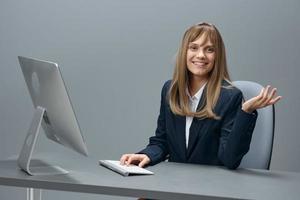 lächelnd tausendjährig blond Geschäftsfrau Arbeiter im Blau Jacke mit Desktop Computer aussehen beim Kamera im grau modern Büro. Fernbedienung Arbeit, Technologie und Werdegang Beruf Konzept. Kopieren Raum foto