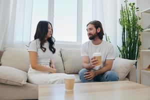 Mann und Frau Sitzung beim Zuhause auf das Couch im Weiß stilvoll T-Shirts Trinken Kaffee aus von Krabbe Tassen von ein Kaffee Geschäft und haben Spaß chatten lächelt und Lachen beim heim. männlich und weiblich Freundschaft foto