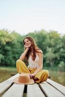 ein jung Frau lächelnd im ein Bild von ein Hippie und Öko-Kleidung Sitzung draußen auf ein Brücke durch ein See tragen ein Hut und Gelb Hose im das Sommer- Sonnenuntergang foto