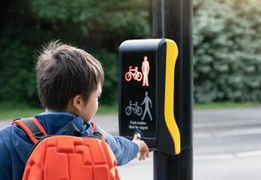 Rückansicht Porträt Schulkind Drücken der Taste an der Ampel auf dem Fußgängerüberweg auf dem Weg zur Schule. Kind Junge mit Rucksack mit Ampel gesteuerten Fußgängereinrichtungen zum Überqueren der Straße foto