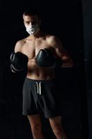 sportlich Mann im medizinisch Maske und im Boxen Handschuhe auf schwarz Hintergrund kurze Hose Fitness Modell- foto