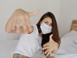 Frau im medizinisch Maske gestikulieren mit ihr Hände während Sitzung auf Bett Pandemie Coronavirus Ferien Quarantäne foto