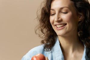 schließen oben Porträt lächelnd lockig schön weiblich im Jeans beiläufig Hemd hält Apfel posieren isoliert auf Über Beige Pastell- Hintergrund. gesund Lebensmittel. natürlich umweltfreundlich Produkte Konzept. Kopieren Raum foto