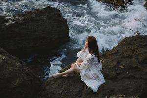 Frau im Weiß Hochzeit Kleid barfuß Sitzung auf ein Stein Natur foto