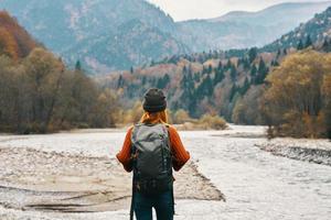 glücklich Frau Wanderer mit ein Rucksack auf das Fluss Bank aussehen in das Berge und Herbst Wald Natur foto