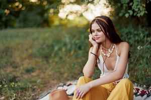 Mädchen gekleidet wie ein Hippie Öko entspannend im das Park, Sitzung auf ein Decke im das Sonnenuntergang, entspannt Lebensstil foto