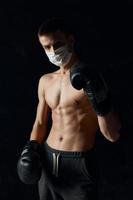 Porträt von ein Boxer im Handschuhe und im grau Töne nackt Torso schwarz Hintergrund medizinisch Maske foto