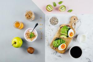 gesund Frühstück Konzept. Sandwich mit Avocado und gekocht Eier, Joghurt mit Granola, Tasse von Kaffee Über dreifarbig stilvoll Hintergrund, oben Sicht, eben legen. Gesundheit, Gewicht Verlust, Diät Essen Konzept foto