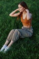 ein schön rothaarig Frau sitzt auf das Grün Gras im ein Garten und sieht aus aus in das Entfernung beim ein Sonnenuntergang umgeben durch Blühen Natur foto