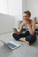 freiberuflich Frau mit Laptop und Telefon funktioniert von Zuhause Sitzung auf das Fußboden im ihr Zuhause Kleider mit ein kurz Haarschnitt, kostenlos Kopieren Raum foto