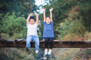 heiter Kinder Sitzung auf hölzern Brücke, glücklich Kinder Junge und Mädchen haben Spaß draussen, Bruder und Schwester spielen im Garten foto