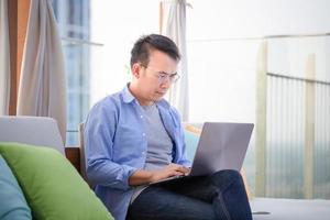 asiatisch Mann mit Laptop Sitzung auf Sofa beim heim, Mann Arbeiten von ein Laptop beim das Terrasse foto
