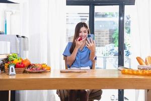 ein jung Frau mit ein schön Gesicht im ein Blau Hemd mit lange Haar Essen Obst Sitzung Innerhalb das Küche beim Zuhause mit ein Laptop und Notizbuch zum Entspannung, Konzept Urlaub. foto