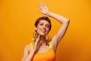 Porträt von ein jung Frau mit ein kurz Haarschnitt und farbig Haar lächelnd im Gelb und zeigen ihr Zunge beim das Kamera auf ein Orange Hintergrund Tanzen mit Ohrringe Zubehör im das Studio foto