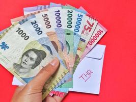 Mann halten Neu Banknoten problematisch im 2022 von rp.1.000 zu Rp. 100.000. indonesisch Rupiah Währung mit ein Weiß Briefumschlag beschriftet thr. Tunjangan Hari raya Konzept isoliert auf rot Hintergrund foto