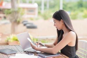 jung Geschäftsfrau schwer beim Arbeit beim ihr Schreibtisch Schreiben Anmerkungen auf ein Pad von ihr Laptop Computer mit analytisch Diagramme und Grafiken im Vorderseite von ihr, hoch Winkel Aussicht foto