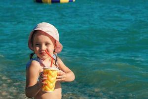 ein wenig Mädchen im ein Baden passen und Panama ist Sitzung auf das Strand, im ihr Hände ist ein Glas mit ein Stroh und ein Sommer- erfrischend Cocktail foto