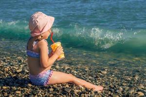 ein wenig Mädchen im ein Baden passen und Panama ist Sitzung auf das Strand, im ihr Hände ist ein Glas mit ein Stroh und ein Sommer- erfrischend Cocktail foto