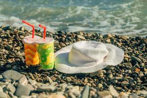 auf das Steine auf das Strand auf ein sonnig Tag Dort sind zwei Cocktails im hell Brille mit Strohhalme, ein Stroh Hut von das Sonne Lügen in der Nähe foto