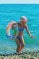 ein wenig Mädchen im ein Baden passen und ein Sonne Hut mit ein aufblasbar Ring läuft von das Welle in das Meer foto