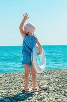 ein wenig Mädchen im Sommer- Strand Kleider und ein Sonne Hut mit ein aufblasbar Ring im ihr Hände steht auf das Strand im Vorderseite von ihr das Blau Meer, Kopieren Raum foto