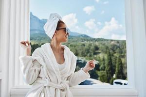 schön Frau tragen Sonnenbrille posieren im ein Bademantel auf ein Balkon sich ausruhen Lebensstil foto