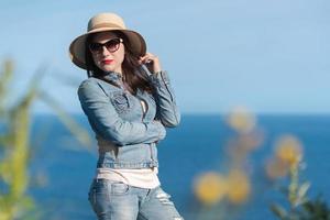 Frau im Sonnenbrille, Stroh Hut, Denim Jacke und Jeans Pose gegen Hintergrund von Blau Himmel und Ozean foto