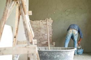 Selektiver Fokus auf Holzhalter im gemischten Zementeimer mit unscharfem Arbeiter und verputztem Zement auf Maurerwand im Hintergrund foto