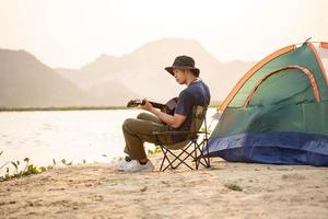 Mann Camping Tourist mit akustisch Gitarre Sitzung auf Sand Strand Singen und spielen Gitarre in der Nähe von touristisch Zelt foto