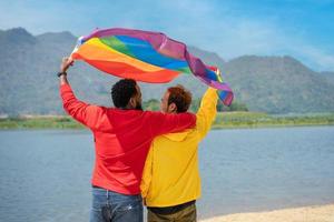 zurück Sicht. afrikanisch Fröhlich und asiatisch Fröhlich Paar Liebe Moment Ausgaben gut Zeit zusammen, erziehen und winken das lgbt Flagge auf das Strand foto