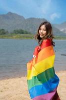 Lesben Frauen mit Fröhlich Stolz Regenbogen Flagge auf das Strand gegen das Himmel. jung Lesben Mädchen halten ein Regenbogen Flagge und umarmen glücklich. Unterstützer von das lgbt Gemeinschaft foto