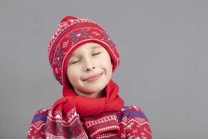 verträumt Kind im warm Kleider auf ein grau Hintergrund. Junge im ein rot gestrickt Hut mit geschlossen Augen. modisch Junge im das Studio auf ein grau Hintergrund. foto