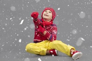 heiter Kind im warm Kleider auf ein Winter Hintergrund. ein Junge im ein rot gestrickt Hut und Pullover. modisch Junge im das Studio im Weiß Schneeflocken foto