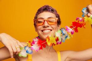 Porträt von ein Frau mit ein Blumen- hawaiisch Kranz um ihr Hals ist haben Spaß Tanzen und lächelnd im hell Kleider auf ein Orange Hintergrund tragen Sonnenbrille, Lebensstil Party hawaiisch Stil foto