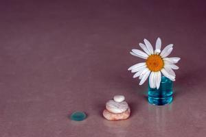 Kamille ist stecken im ein Flasche mit Blau aromatisch Öl und drei pyramidenförmig Kieselsteine und ein Glas Stück auf ein Rose Hintergrund von Keramik Fliesen. selektiv Fokus auf das Kiesel. Platz zum Text. foto