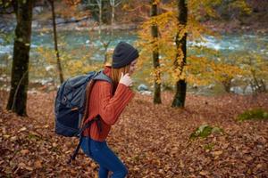 Frau im ein Sweatshirt mit ein Rucksack auf ihr zurück in der Nähe von das Fluss im das Berge und Park Bäume Herbst Landschaft foto