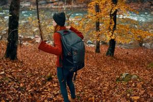 Frau im ein Sweatshirt mit ein Rucksack und im Jeans Spaziergänge durch das Herbst Wald im das Berge in der Nähe von das Fluss foto