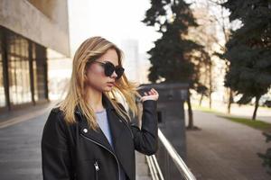 modisch Frau Brille Modell- Leder Jacke Sonne Park Gebäude foto