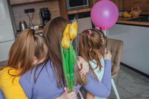 Töchter gratulieren ihr Mama auf Mutter Tag, ein Karte mit ein Herz, Blumen und ein Ballon beim Zuhause im das Küche, Sie alle Umarmung. Kinder Überraschung ihr Mutter zum das Urlaub. foto